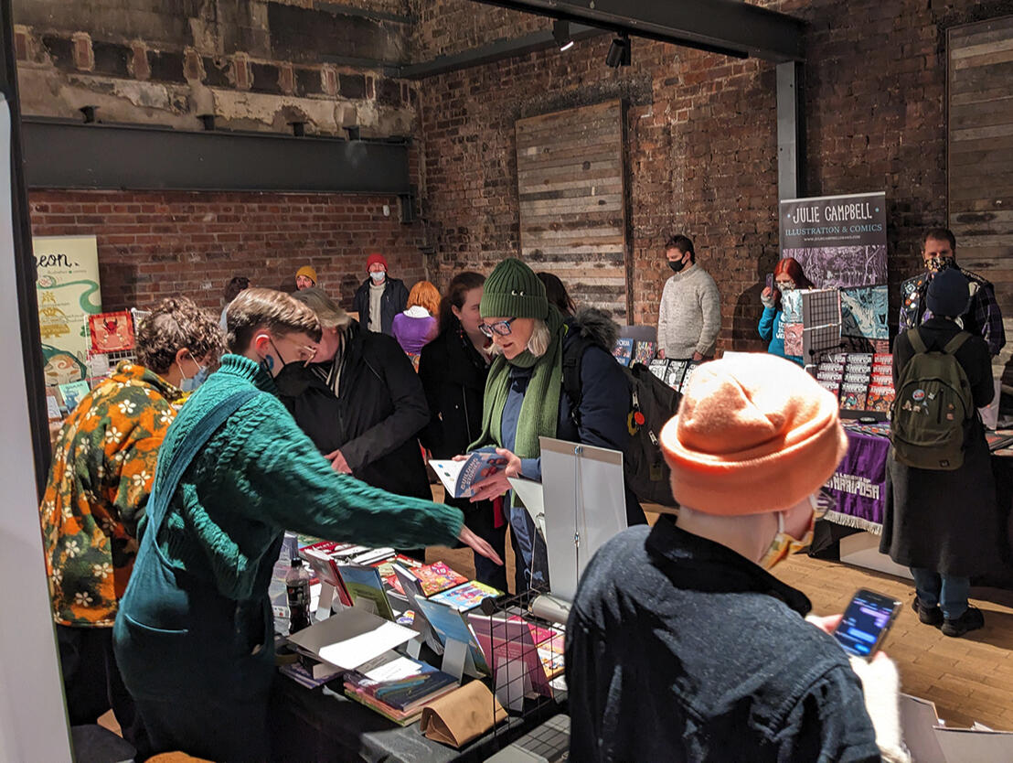 Photo from a behind a vendors table at TAGS 23. An exhibitor is talking to a visitor about various books on their table. All exhibitors are wearing masks, and some guests are wearing masks.