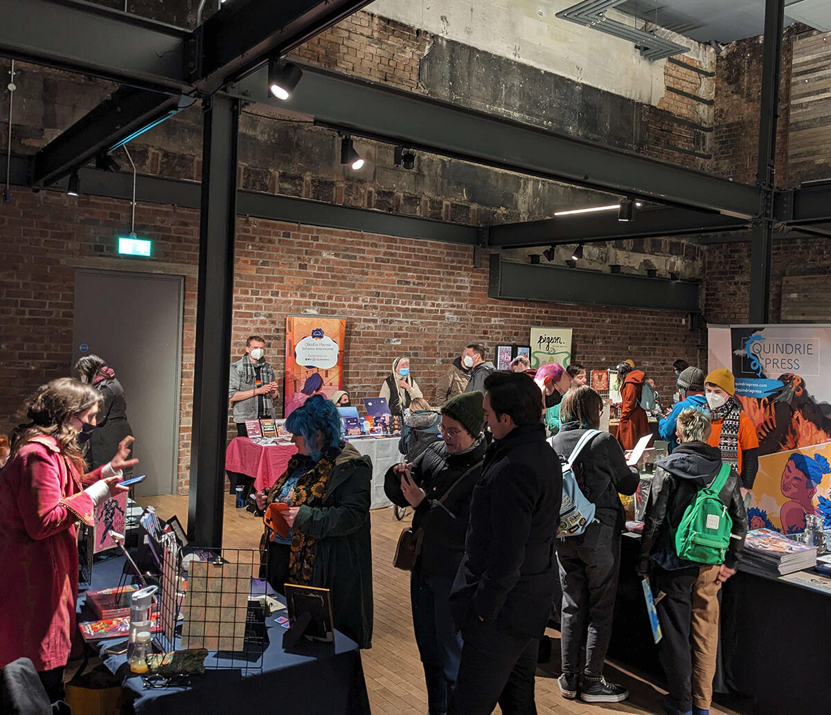 Photo from TAGS 23, showing a busy indoor marketplace. Many exhibitors and some visitors are wearing masks.