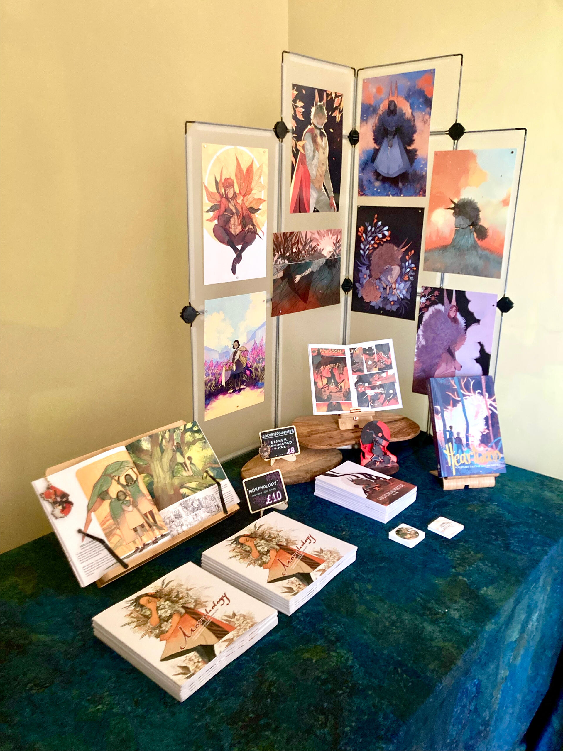 Photo of a convention table set up. On the table are two stacks of books, accompanied with spread open displays of those books on plinths. On one corner of the table is a plastic grid frame, with prints attached.